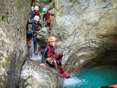 Eine Gruppe von Kindern in Neoprenanzügen und Helmen klettert über glatte Felsen in einem klaren, blauen Wasserbecken.