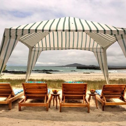 Unter einem gestreiften Sonnenschirm stehen mehrere Holzliegestühle am Strand, mit Blick auf das ruhige Wasser und die Küste.