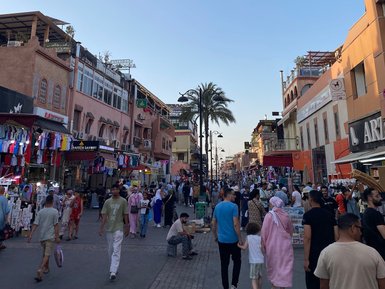 Belebte Straße in Marrakesch voller Menschen, farbenfrohe Stände und Schilder, typische Atmosphäre des Medinas.