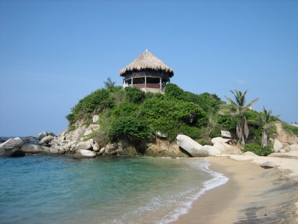 Ein malerisches Strandhaus mit Strohdach thront auf einer bewachsenen Klippe, umgeben von klarem Wasser und sanften Wellen.