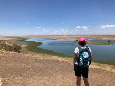 Eine Person mit einem Rucksack steht am Ufer eines ruhigen Gewässers und blickt auf die weite Landschaft von Amboseli.