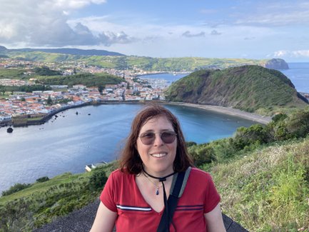 Eine Frau mit Sonnenbrille lächelt vor einer malerischen Küstenlandschaft mit Bergen und einem ruhigen Meer.