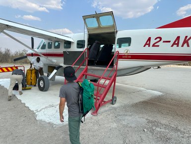 Ein kleiner Flugzeug steht auf einem staubigen Platz, während Passagiere mit Gepäck einsteigen und aussteigen.