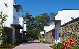 Eine sonnige Straße zwischen modernen, weißen Ferienhäusern, umgeben von üppigem Grün und bunten Pflanzen.