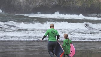 Eine Frau und ein Mädchen in grünen Neoprenanzügen gehen mit Bodyboards am Strand entlang, während die Wellen brechen.