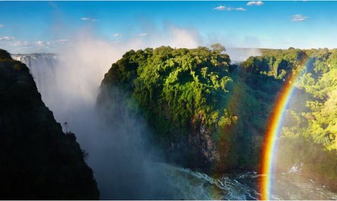 Ein atemberaubender Blick auf die Victoriafälle, umgeben von üppigem Grün und einem strahlenden Regenbogen im Hintergrund.