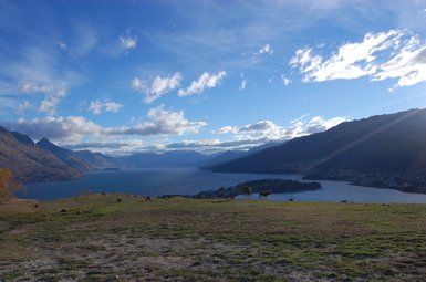 Eine weite Landschaft mit sanften Hügeln, einem glitzernden See und majestätischen Bergen unter einem strahlend blauen Himmel.