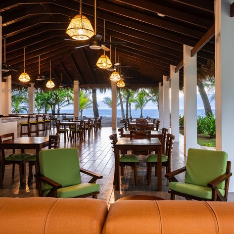Ein einladendes Restaurant mit Holztischen und Stühlen, umgeben von Palmen und Blick auf das Meer.