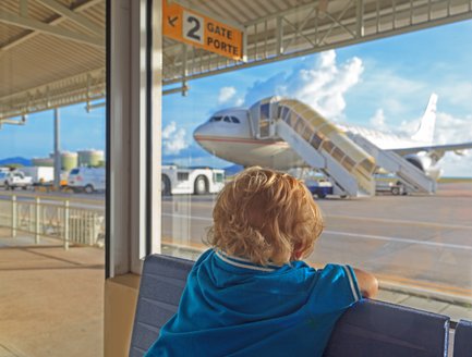 Ein kleines Kind mit blonden Locken sitzt am Fenster und beobachtet ein Flugzeug, das am Gate parkt.