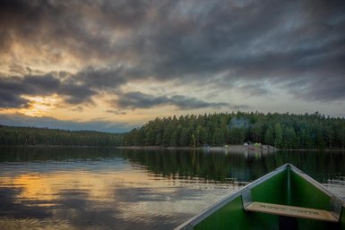 Ein ruhiger See spiegelt die Wolken und Bäume wider, während die Abendsonne hinter dem Horizont verschwindet.