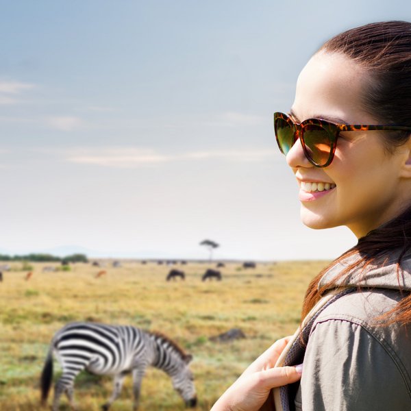 Eine lächelnde Frau mit Sonnenbrille steht in einer weiten Savanne, während Zebras im Hintergrund grasen.