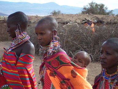 Drei Frauen in traditioneller Maasai-Kleidung stehen nebeneinander, eine trägt ein Baby auf dem Rücken.