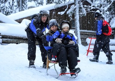 Eine Familie genießt eine Schlittenfahrt im verschneiten Wald, während Schneeflocken sanft fallen und die Umgebung verzaubern.