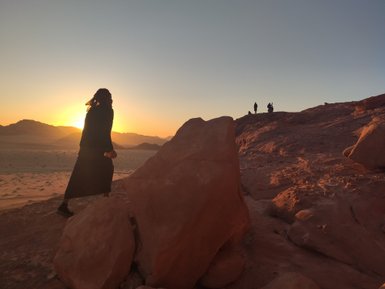 Eine Person in schwarzer Kleidung wandert über felsiges Terrain, während die Sonne hinter den Bergen untergeht.