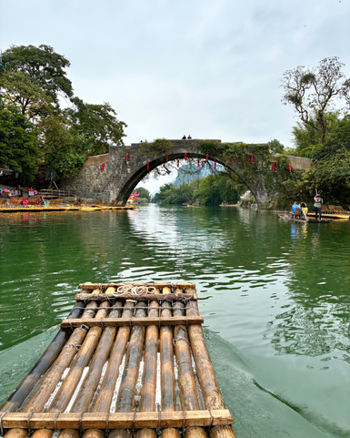 Ein Bambusfloß schwimmt ruhig auf einem klaren Fluss, während ein gewölbter Steinbogen im Hintergrund sichtbar ist.