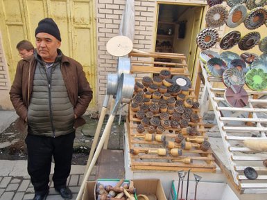 Ein Mann steht an einem Marktstand, umgeben von handgefertigten Holzgegenständen und bunten Schalen im Hintergrund.