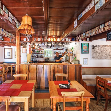 Ein gemütliches Restaurant mit Holzmöbeln, einem Barkeeper, der Getränke hinter der Theke serviert, und bunten Wanddekorationen.