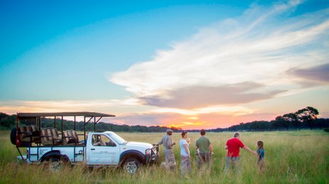 Eine Gruppe von fünf Personen steht in einem weiten Grasfeld, während die Sonne am Horizont untergeht.