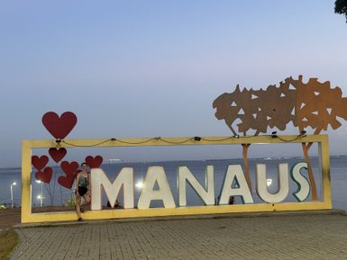 Eine Person lehnt entspannt an einem bunten 'Manaus'-Schild, umgeben von herzförmigen Dekorationen und einer ruhigen Wasserlandschaft.