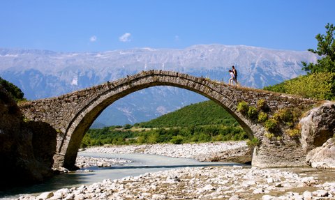 Ein Paar geht über eine alte Steinbrücke, umgeben von grünen Hügeln und majestätischen Bergen unter einem strahlend blauen Himmel.
