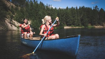 Ein Paar paddelt fröhlich in einem blauen Kanu auf einem ruhigen Fluss, umgeben von üppigem Wald und klarem Himmel.