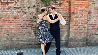 Ein Paar tanzt leidenschaftlich Tango vor einer roten Ziegelmauer, umgeben von einer lebhaften Atmosphäre.