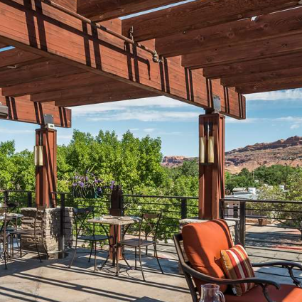 Eine sonnige Terrasse mit Holzpergola bietet einen Blick auf grüne Bäume und rote Felsen im Hintergrund.