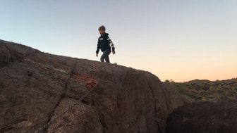 Ein Junge klettert auf einen großen Felsen, während der Himmel in sanften Farben des Sonnenuntergangs leuchtet.