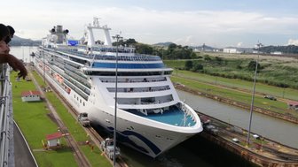 Ein großes Kreuzfahrtschiff passiert die Miraflores-Schleuse im Panamakanal, umgeben von grüner Landschaft und Zuschauern.