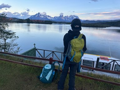 Ein Reisender mit einem grünen Rucksack steht am Ufer eines ruhigen Gewässers und blickt auf majestätische Berge.
