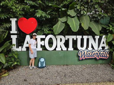 Eine Person steht lächelnd neben einem großen Schild, das 'I LOVE LA FORTUNA' und einen Wasserfall zeigt, umgeben von üppigem Grün.