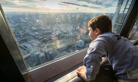 Ein Junge schaut fasziniert aus einem hohen Fenster auf die Stadt Toronto, während die Sonne am Horizont untergeht.