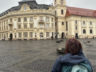Eine Person mit einem Rucksack steht auf einem Platz und blickt auf ein historisches Gebäude mit einer markanten Uhr.