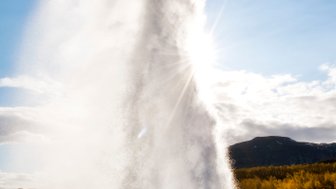 Ein beeindruckender Geysir sprudelt mit kraftvollem Wasserstrahl in den klaren blauen Himmel, umgeben von sanften Hügeln.