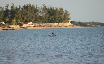 Ein Mann paddelt in einem kleinen Boot auf ruhigem Wasser, während im Hintergrund eine Küstenlandschaft sichtbar ist.