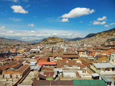 Ein weitläufiger Blick auf die Stadt mit zahlreichen Dächern und einem Hügel im Hintergrund unter einem strahlend blauen Himmel.