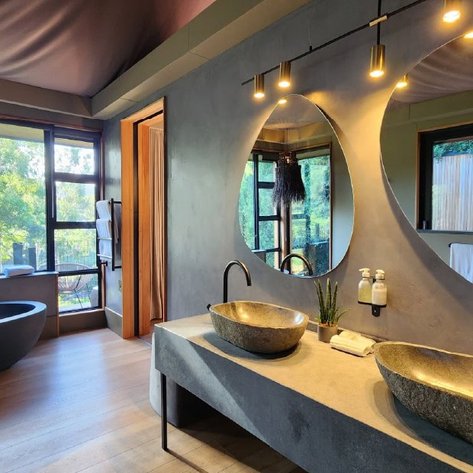 Ein modernes Badezimmer mit zwei eleganten Waschbecken aus Stein und großen Spiegeln, umgeben von natürlichem Licht.