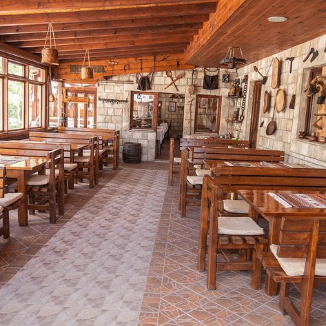 Ein rustikales Restaurant mit Holztischen und Stühlen, umgeben von Steinwänden und großen Fenstern, die Tageslicht hereinlassen.