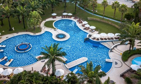 Ein weitläufiger Pool mit blauen Fliesen, umgeben von Palmen und Liegen, lädt zum Entspannen in der Sonne ein.