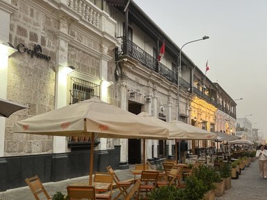 Eine belebte Straße in Arequipa mit Cafés, Holzstühlen und Tischen unter Sonnenschirmen, umgeben von blühenden Pflanzen.