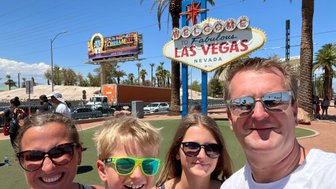 Eine Familie posiert fröhlich vor dem berühmten Las Vegas-Schild, umgeben von Palmen und strahlendem Sonnenschein.