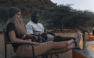 Eine Frau mit lockigem Haar und ein Mann sitzen entspannt am Lagerfeuer, während sie Getränke in der Hand halten.