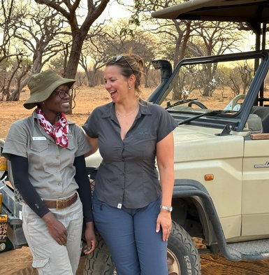 Zwei Frauen stehen lächelnd neben einem Jeep in der afrikanischen Savanne, umgeben von trockenen Bäumen und einem warmen Licht.