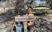 Zwei Jungen stehen neben einem Schild mit der Aufschrift 'YOUNG EXPLORERS' in einer sonnigen, bewaldeten Umgebung.