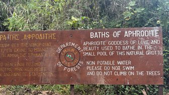 Ein Holzschild beschreibt die Bäder der Aphrodite, umgeben von üppigem Grün und einem kleinen natürlichen Pool.