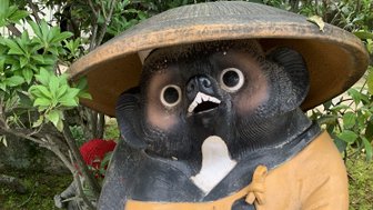 Eine große, freundliche Statue eines Tanuki mit einem Hut, die eine Schüssel in den Händen hält, umgeben von Pflanzen.