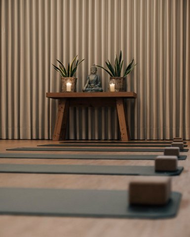 Ein ruhiger Yogaraum mit mehreren Matten, einem Buddha-Statuen und Pflanzen auf einem Holztisch, umgeben von sanftem Licht.