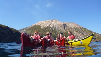 Sechs Kajakfahrer paddeln in ruhigem Wasser, während ein majestätischer Vulkan im Hintergrund thront.