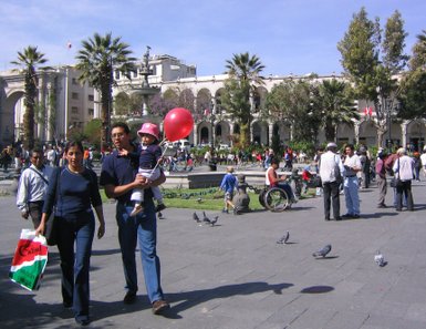 Eine Familie spaziert durch den Plaza de Armas, während ein Kind einen roten Ballon hält und die Sonne scheint.