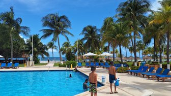 Zwei Jungen in Badehosen gehen am Pool entlang, umgeben von Palmen und Liegen unter einem strahlend blauen Himmel.